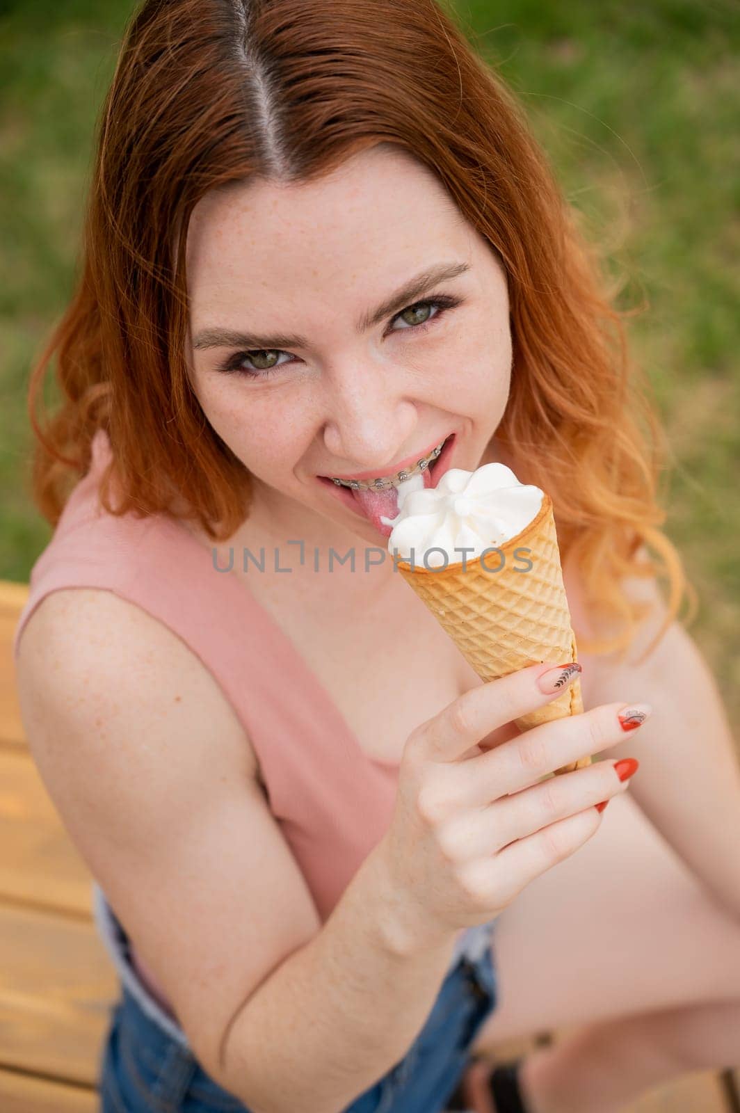 Young beautiful red-haired woman smiling with braces and eating an ice cream cone outdoors. Vertical photo. by mrwed54