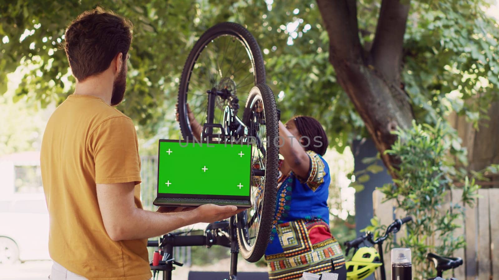 Young man with laptop displaying greenscreen as african american lady attaches wheel to bicycle frame in yard. Personal computer with isolated chromakey template held by caucasian male.