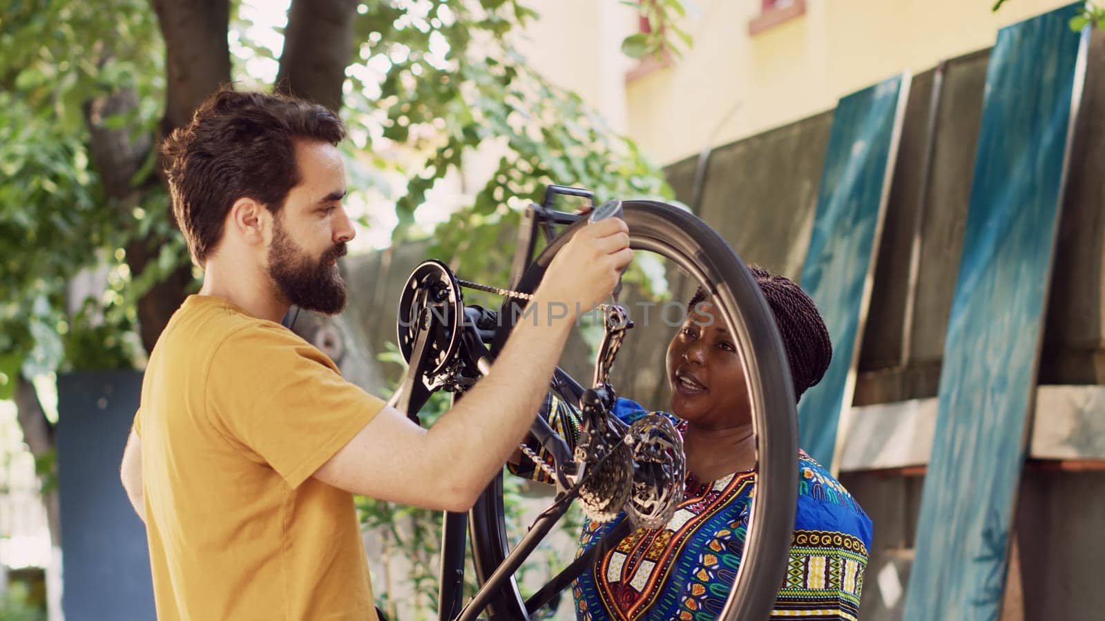 Sporty multiethnic couple doing bike maintenance in home yard. Young african american woman grasping modern bicycle while healthy caucasian man diligently lubricating wheels outdoor.