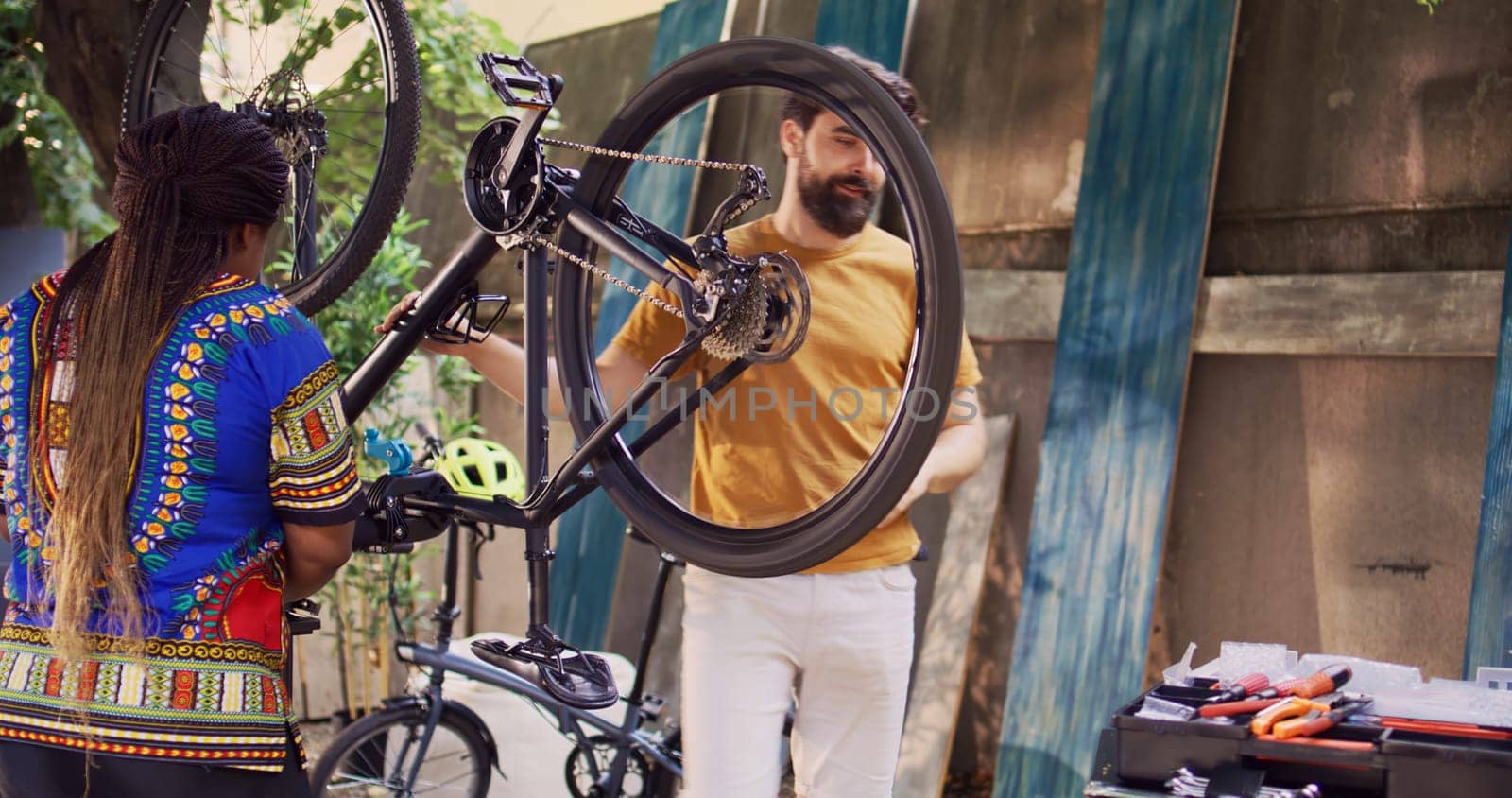 Young sports-loving multiracial couple repairing broken bicycle using professional equipment. Caucasian male loosening and dismantling damaged bike tire while black woman prepares work tools.