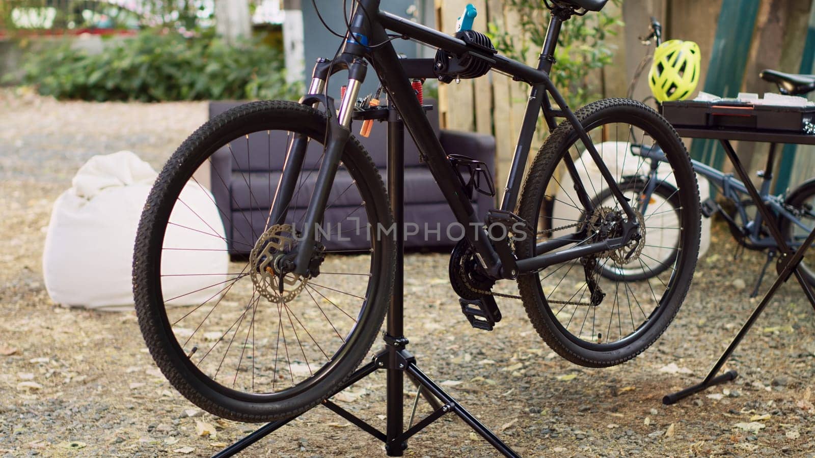 Modern bicycle is visible in the yard positioned on repair-stand for examining and mending. Outdoor various bike components await adjustments and restoration with professional work tools.