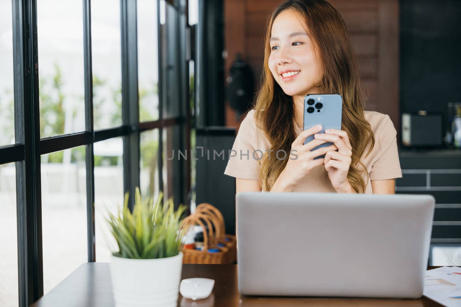 Happy lifestyle female smiling using smartphone in coffee shop, young business woman working with laptop computer she holding smart mobile phone looking out of windows at cefa for texting messages