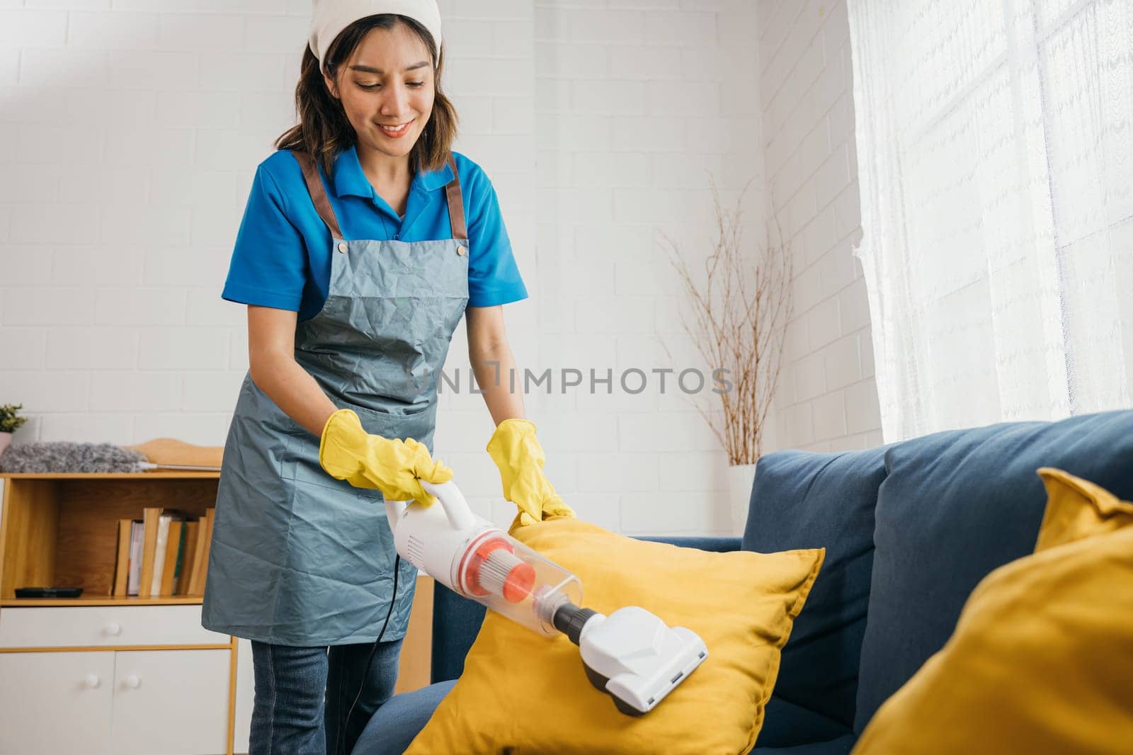 Focused on housework an Asian woman maid cheerfully vacuum machine cleaners a sofa in a living room. Her dedication to hygiene and furniture care reflects modern cleaning approaches.