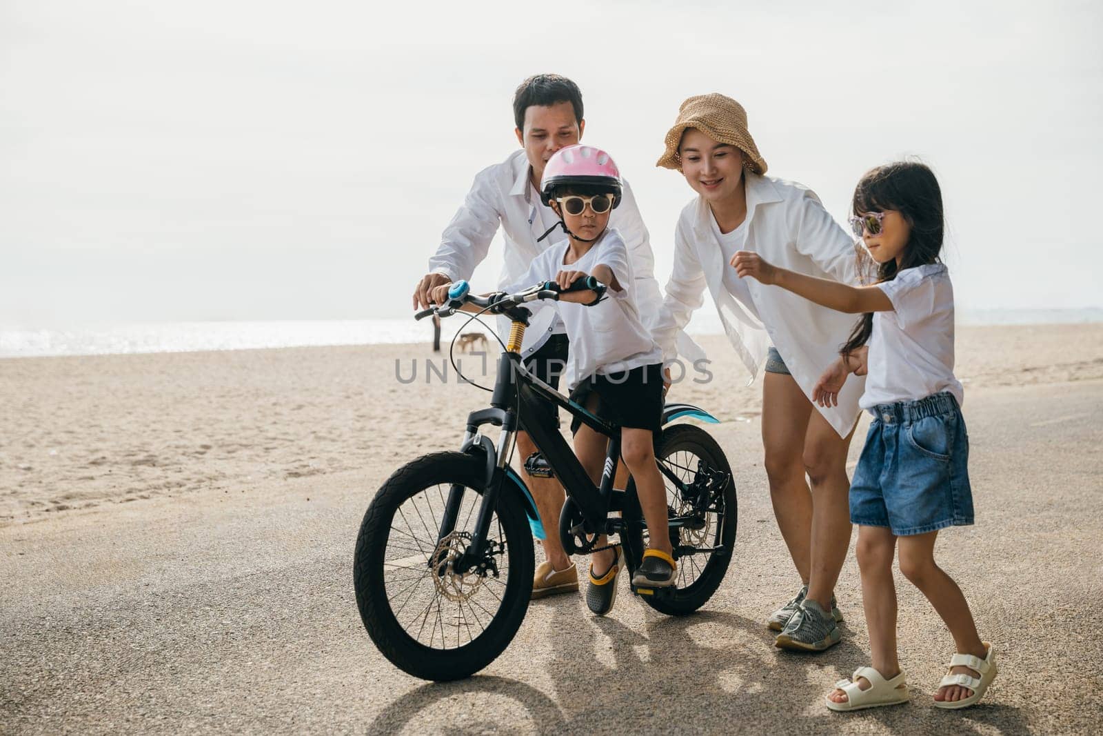 Family fun under summer sun: Parents happily teach their children to ride bicycles on sandy beach near sea. Laughter safety helmets and freedom of cycling make this joyful and memorable moment.