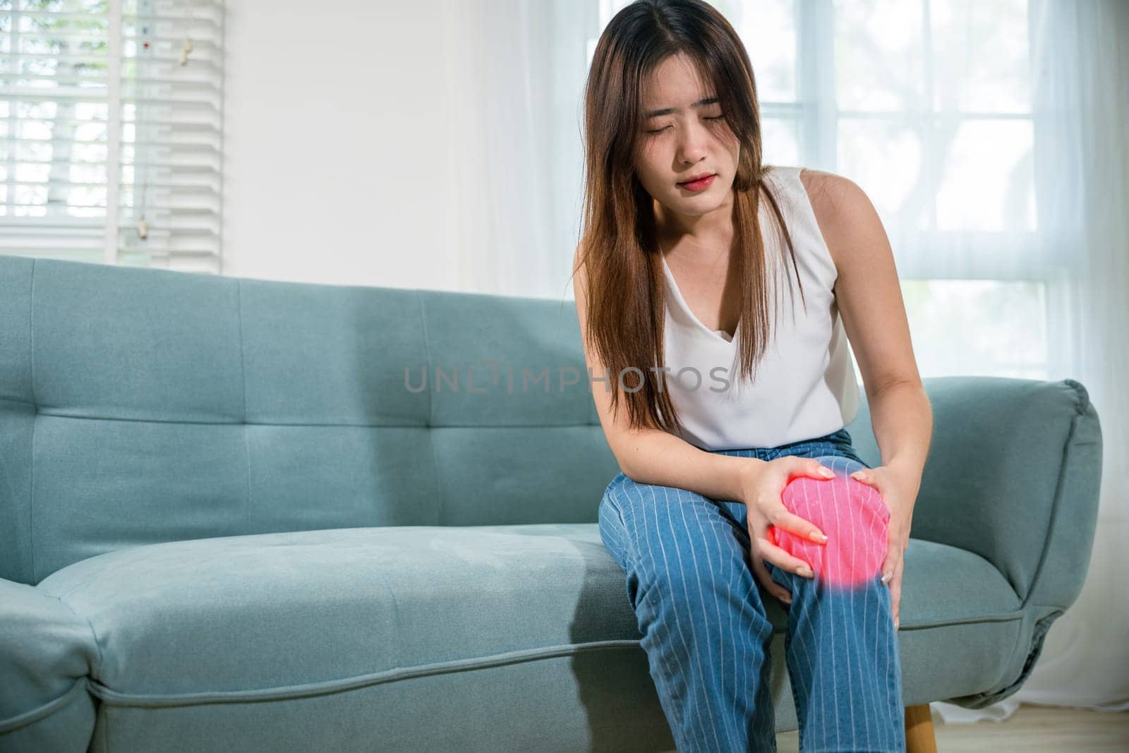 Asian young woman sitting on sofa and feeling suffering knee pain by Sorapop