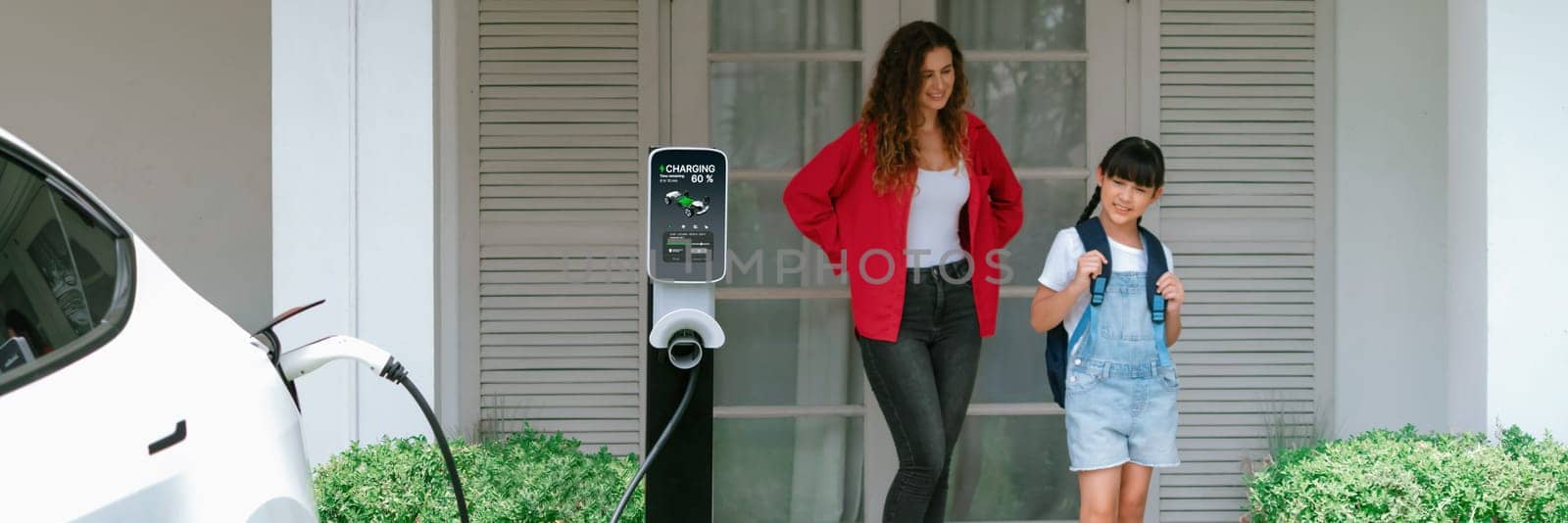 Happy little young girl learn about eco-friendly and energy sustainability as she help her mother recharge electric vehicle from home EV charging station. EV car and modern family. Panorama Synchronos