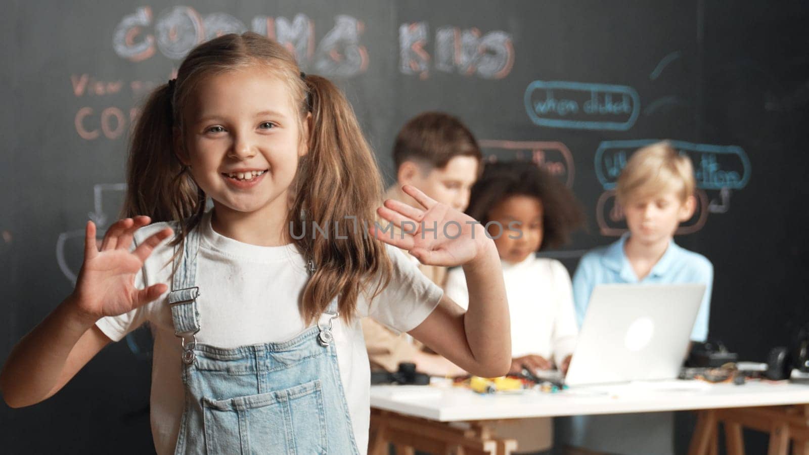 Girl smile and wave to camera while friend working or learning engineering code or prompt in STEM technology classroom. Student looking at camera and greeting while children using laptop. Erudition.