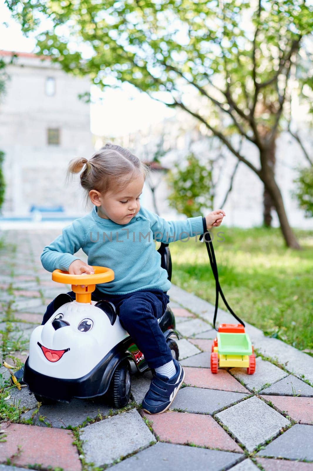 Little girl sitting on a toy car with a small toy car on a rope on a path in the garden. High quality photo