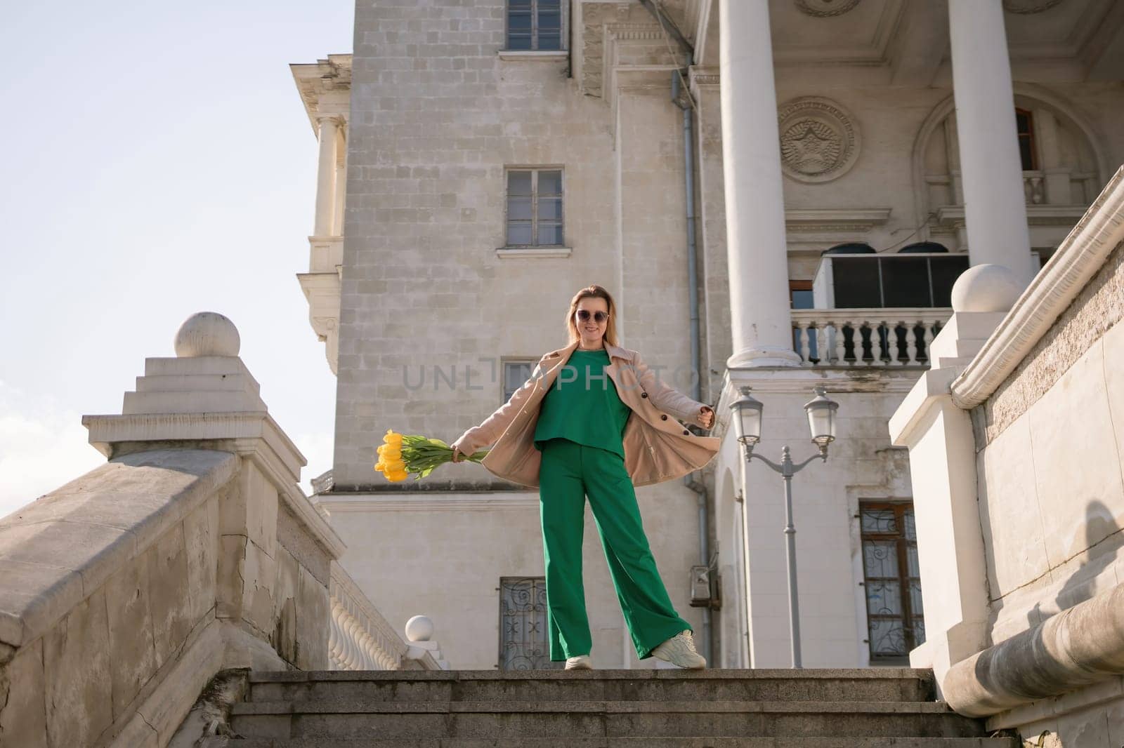 A woman in a green outfit stands on a set of stairs in front of a building. She is holding a bouquet of yellow flowers. The scene has a sense of elegance and sophistication