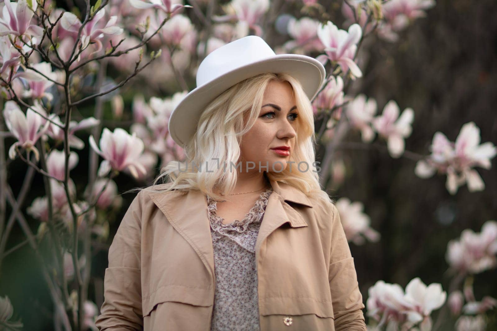 A blonde woman wearing a white hat stands in front of a tree with pink flowers by Matiunina