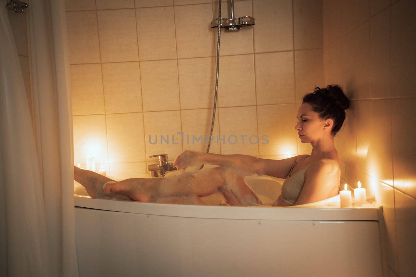 A woman is sitting in a bathtub with candles lit around her. Scene is relaxing and calming