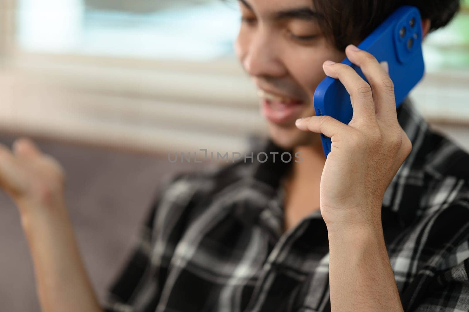 Smiling young Asian man having phone conversation while sitting on couch at home.