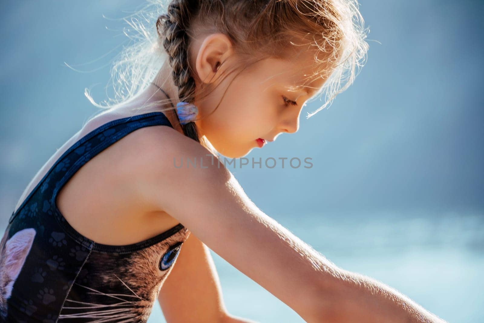 A young girl is sitting on the beach, looking at the water. She is wearing a blue tank top with a cat on it