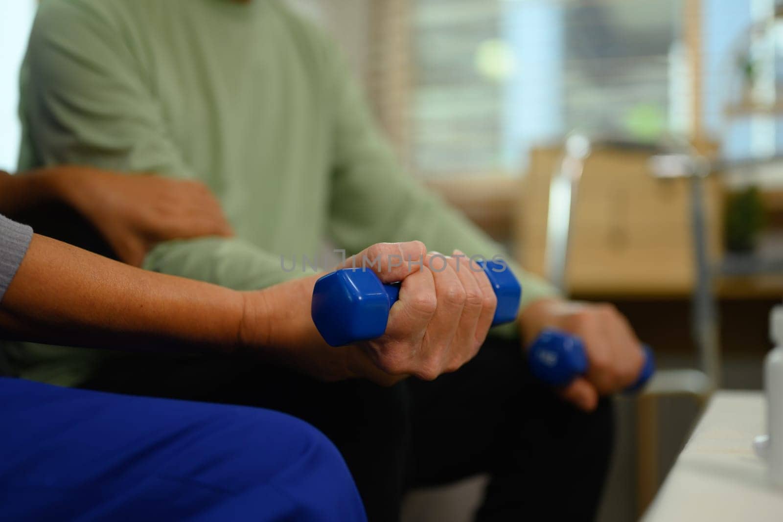Nurse or physiotherapist caregiver helping senior man exercising with dumbbell at home.