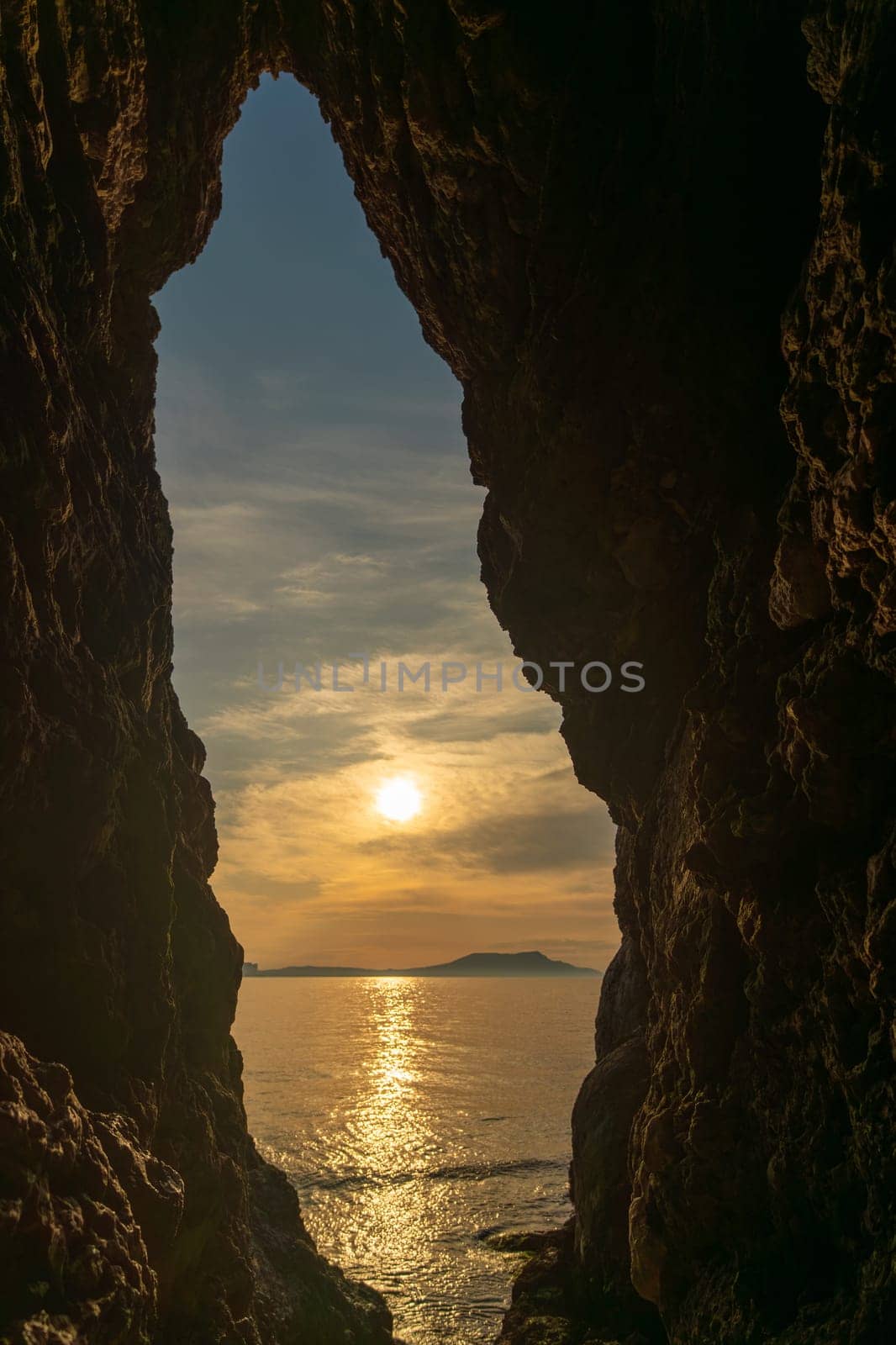 A large hole in a rock wall with the sun shining through it. The sun is setting and the sky is cloudy. by Matiunina