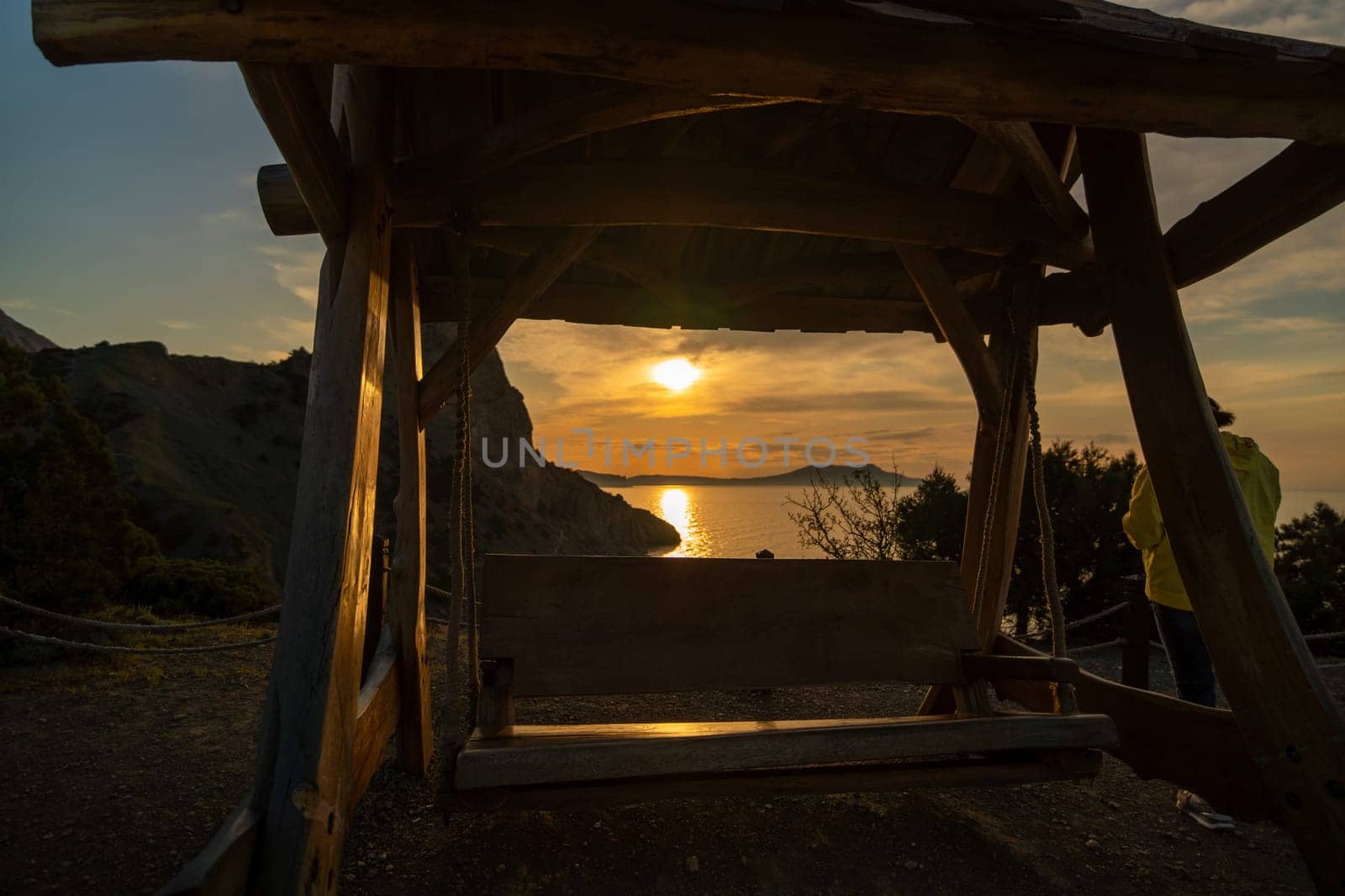 A wooden swing is suspended over a rocky shoreline. The sun is setting, casting a warm glow over the scene. by Matiunina