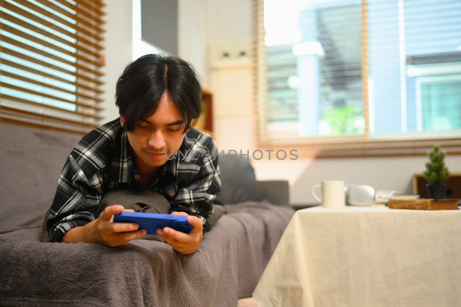 Relaxed young Asian man lying on sofa and checking social media on smartphone by prathanchorruangsak