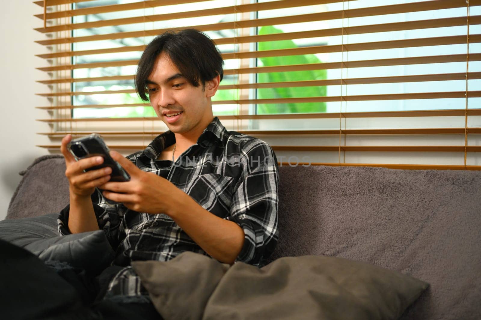 Happy young man using mobile phone sitting on couch at home by prathanchorruangsak