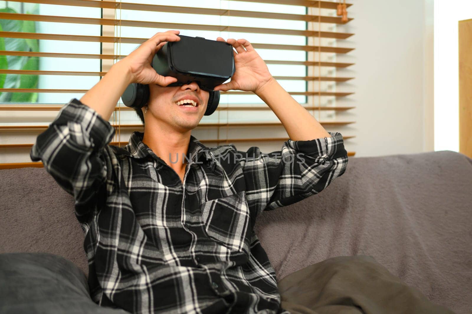 Smiling man wearing VR headset sitting on couch, spending weekend at home by prathanchorruangsak