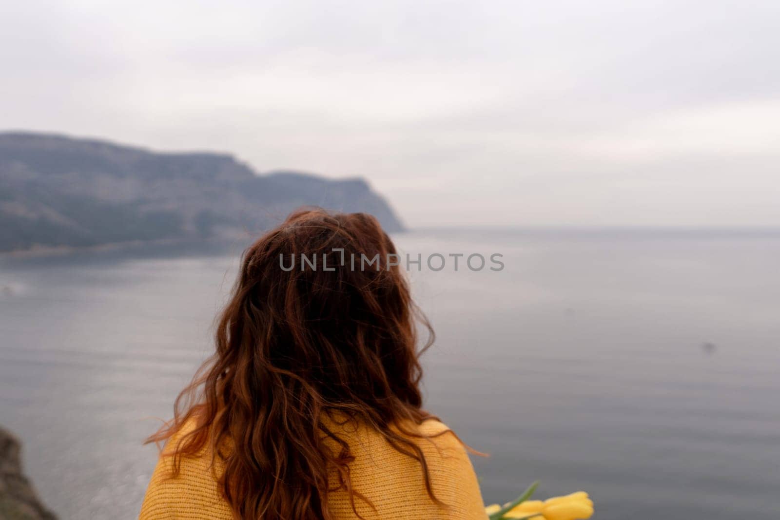 Rear view of a woman with long hair against a background of mountains and sea. Holding a bouquet of yellow tulips in her hands, wearing a yellow sweater.