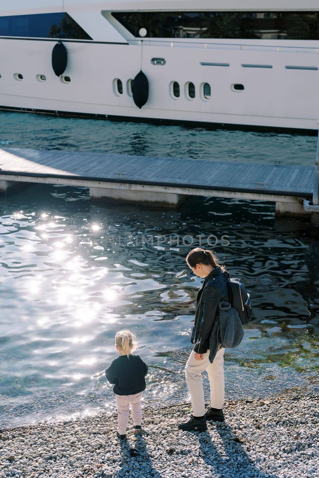 Mother and a little girl stand on a pebble beach near a moored yacht and look at the water. Back view by Nadtochiy