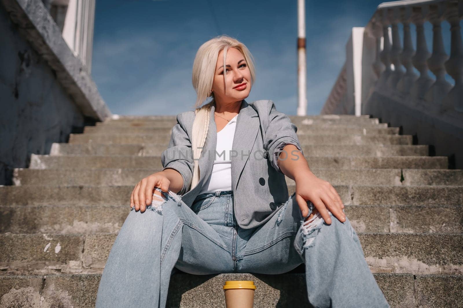 A woman in a gray jacket and blue jeans sits on a set of stairs. She is smiling and she is enjoying herself