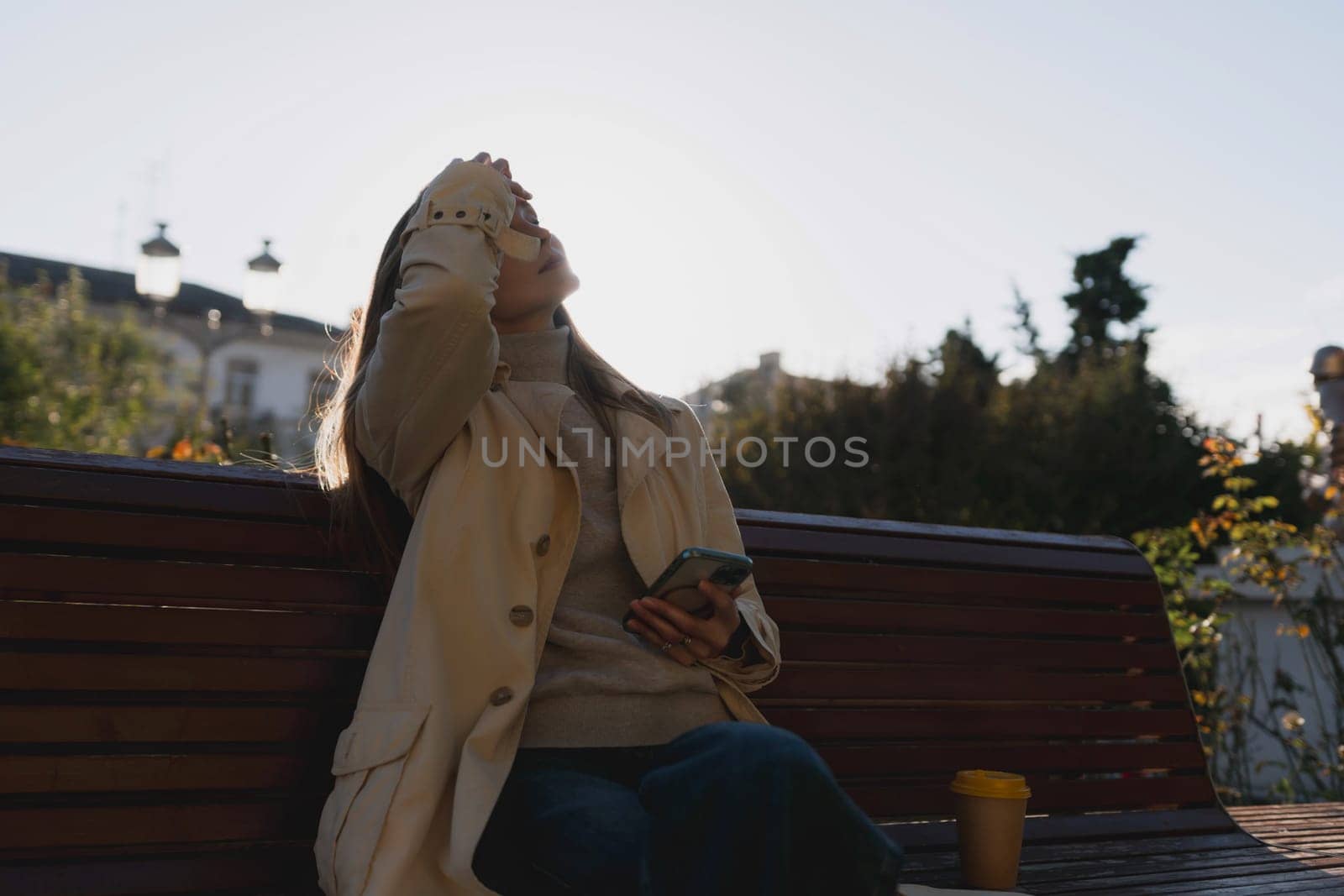 A woman sitting on a bench with her hand on her face. She is wearing a tan coat and holding a cell phone