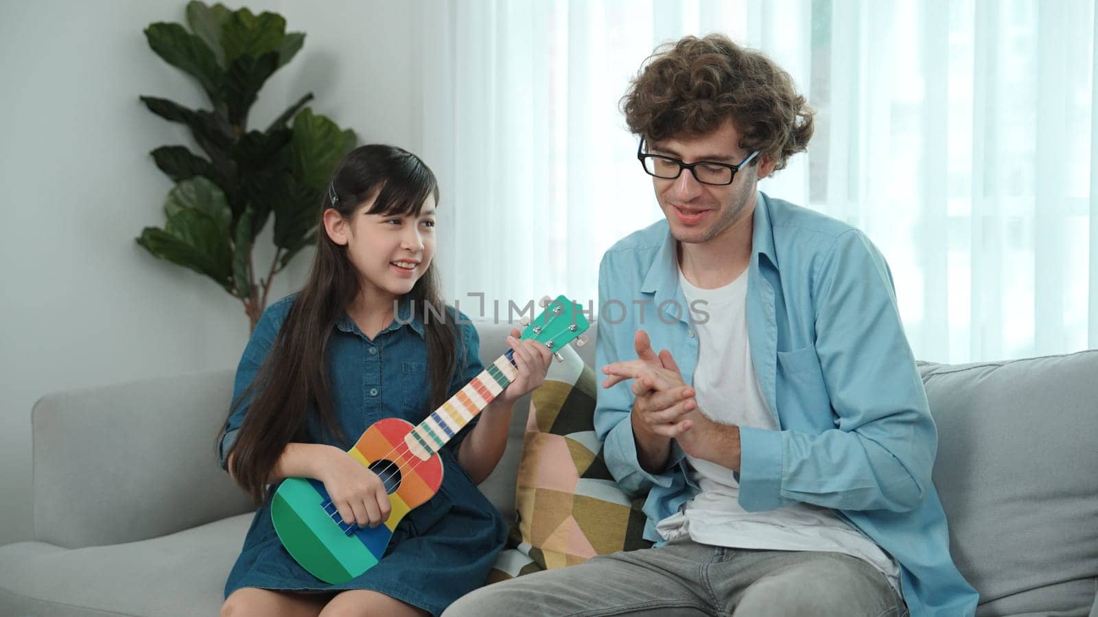 Back view of happy father looking at smart daughter playing ukulele warmly and singing song. Happy dad motivated and encouraged american school girl enjoy playing guitar. Family recreation. Pedagogy.