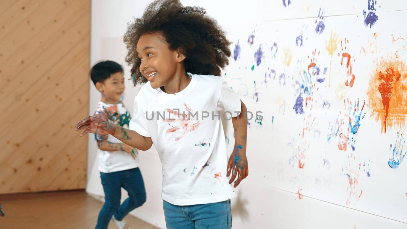 Young asian boy running to stamp colored hand print at cute african girl shirt. Student playing in art lesson with white background stained with colorful color. Creativity activity concept. Erudition.