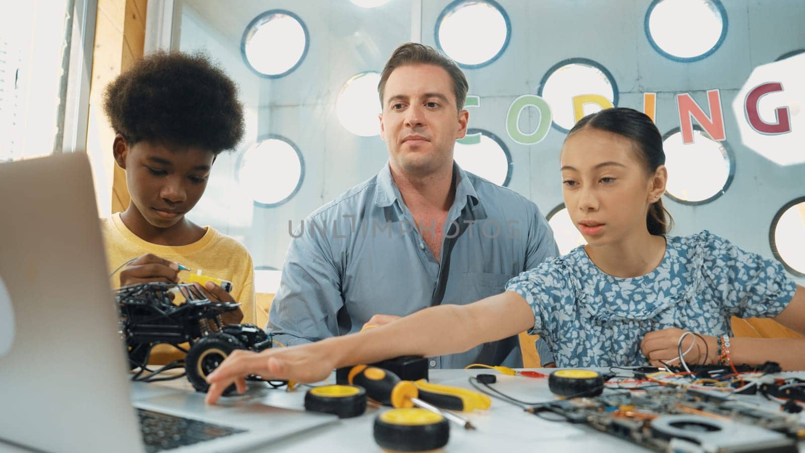 Caucasian student asking teacher about code and pointing at laptop screen. Professional instructor answer girl question while boy repair car model at table with electronic tool placed. Edification.