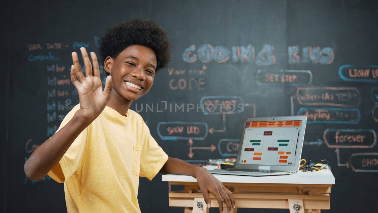 Smart african boy use laptop to code program and turn around to wave hand. Cute highschool stident coding engineering prompt or program system at blackboard in STEM technology classroom. Edification