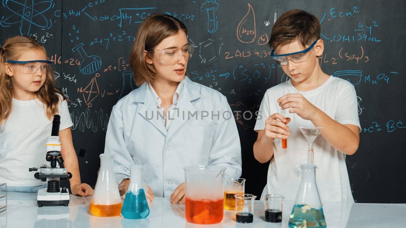 Caucasian boy mixing chemical liquid while teacher giving advice. Professional instructor wearing lab suit looking for diverse student at table with beaker filled with colored solution. Erudition.