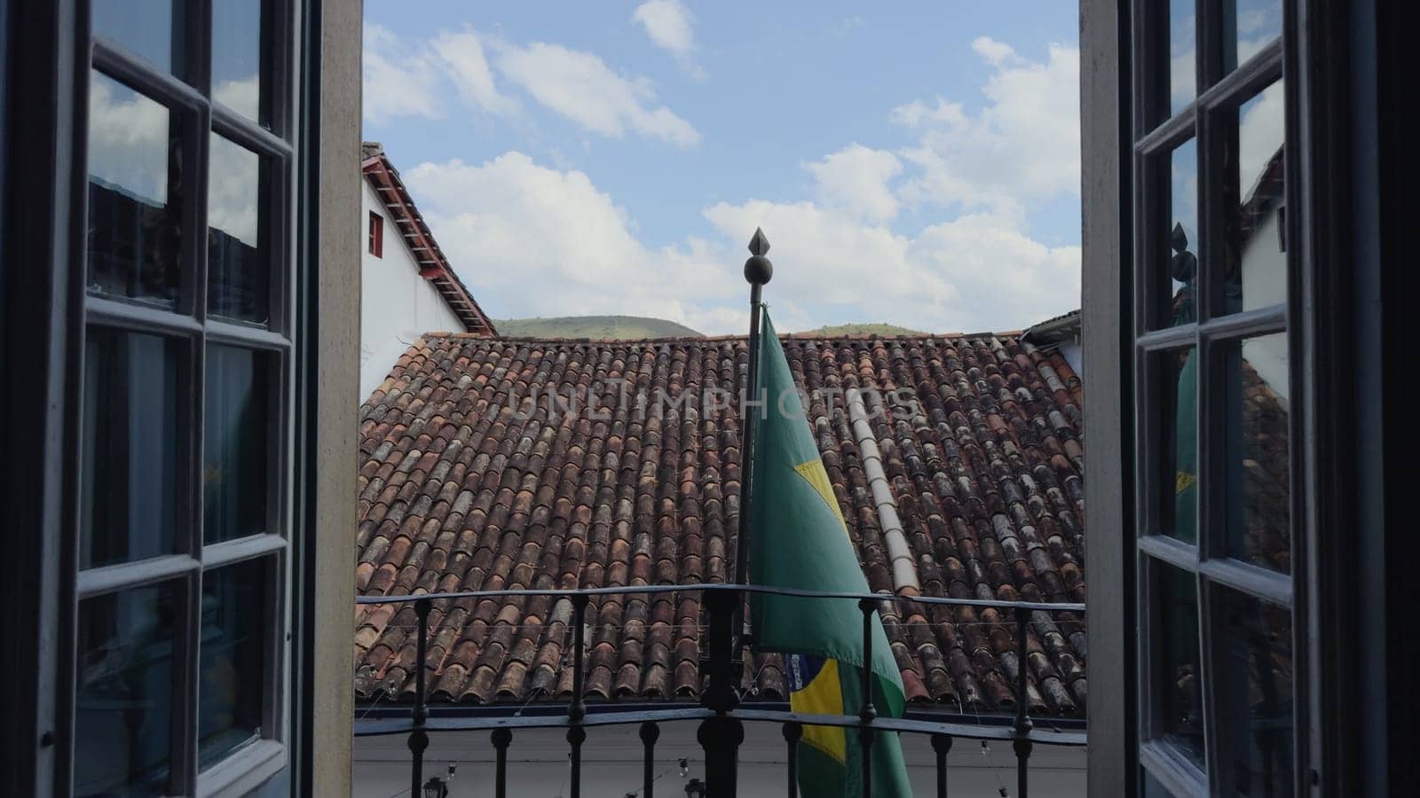 Brazilian flag on a balcony with open doors, building's roof in the background.