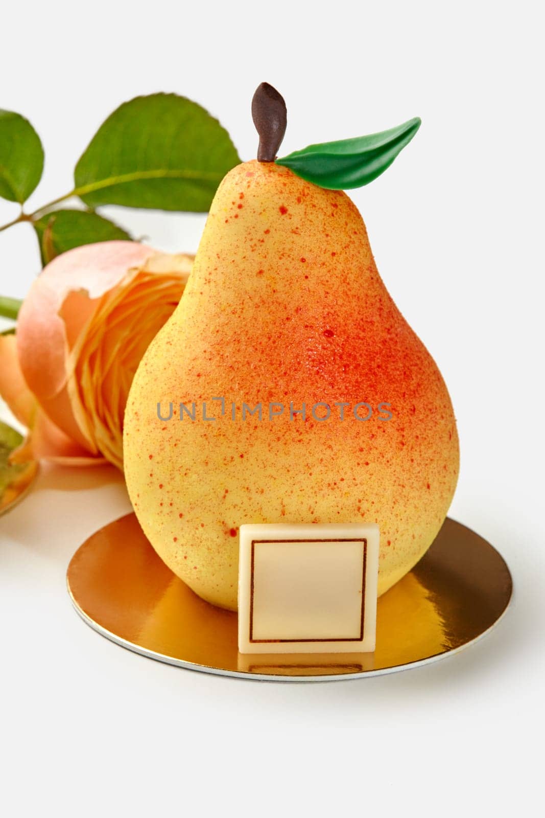 Appetizing pear-shaped mousse dessert made with fruit confit decorated with colored chocolate leaf and stem on golden serving cardboard on white background with fresh rose