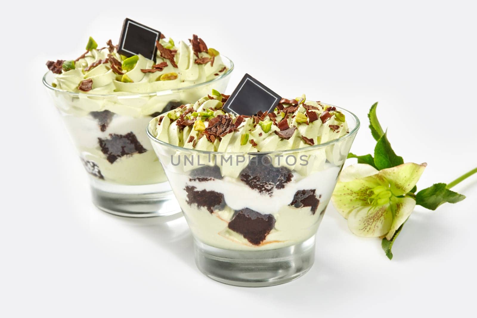 Luxurious pistachio sundae with pieces of brownie and whipped cream, topped with chocolate shavings and nuts served in clear dessert glasses on white background with delicate flower