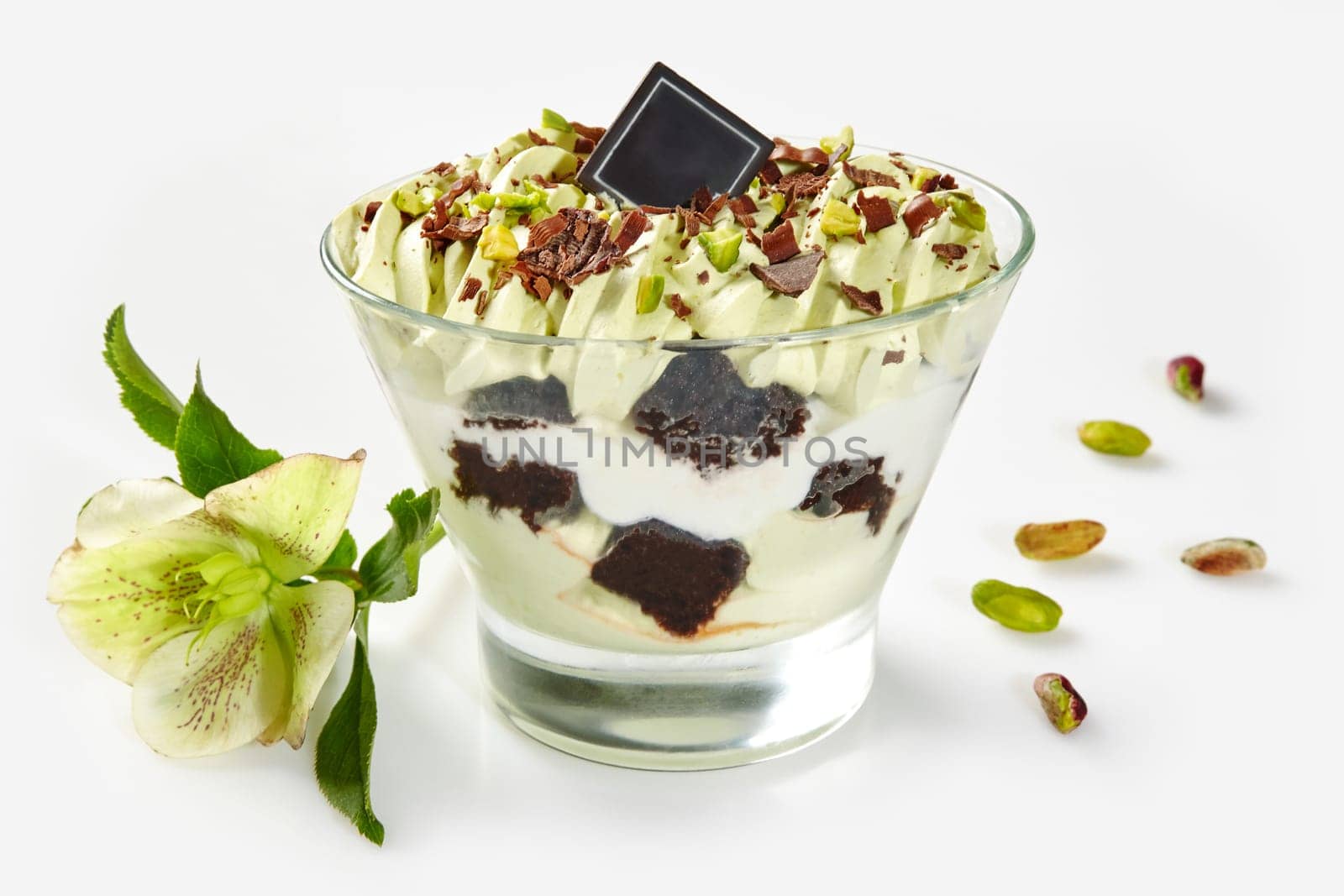 Exquisite pistachio-flavored layered dessert garnished with chocolate and pistachio nuts, accompanied by delicate greenish flower on white background