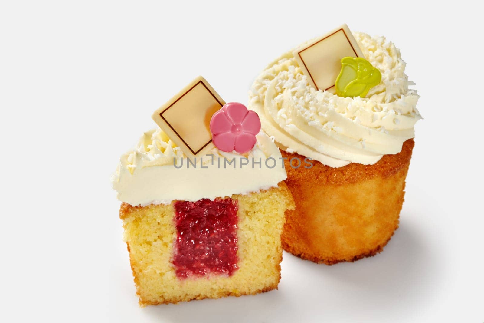 Appetizing whole and sliced fluffy vanilla cupcakes with raspberry filling topped with swirls of whipped cream sprinkled with coconut shavings and chocolate plates. Popular baked desserts