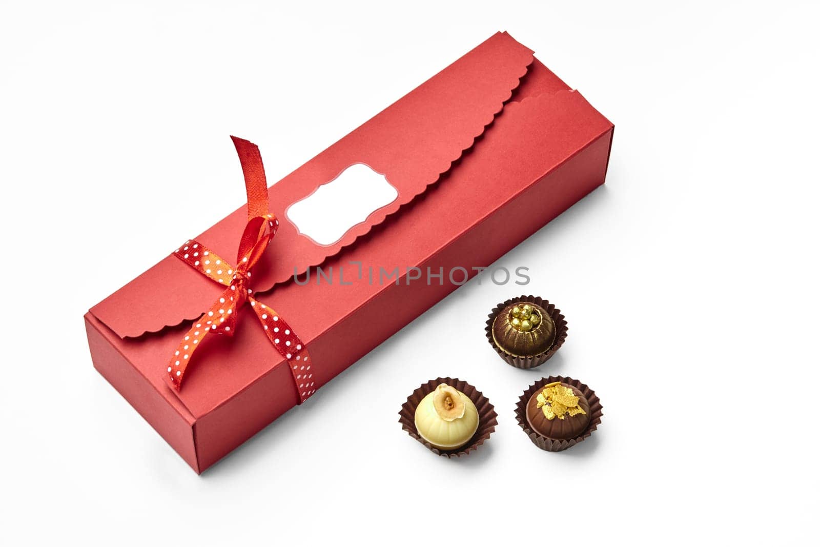 Artisan chocolate candies decorated with nuts, caramel shavings and golden sugar pearls encased in elegant red box with signature label tied with dotted ribbon for gifting