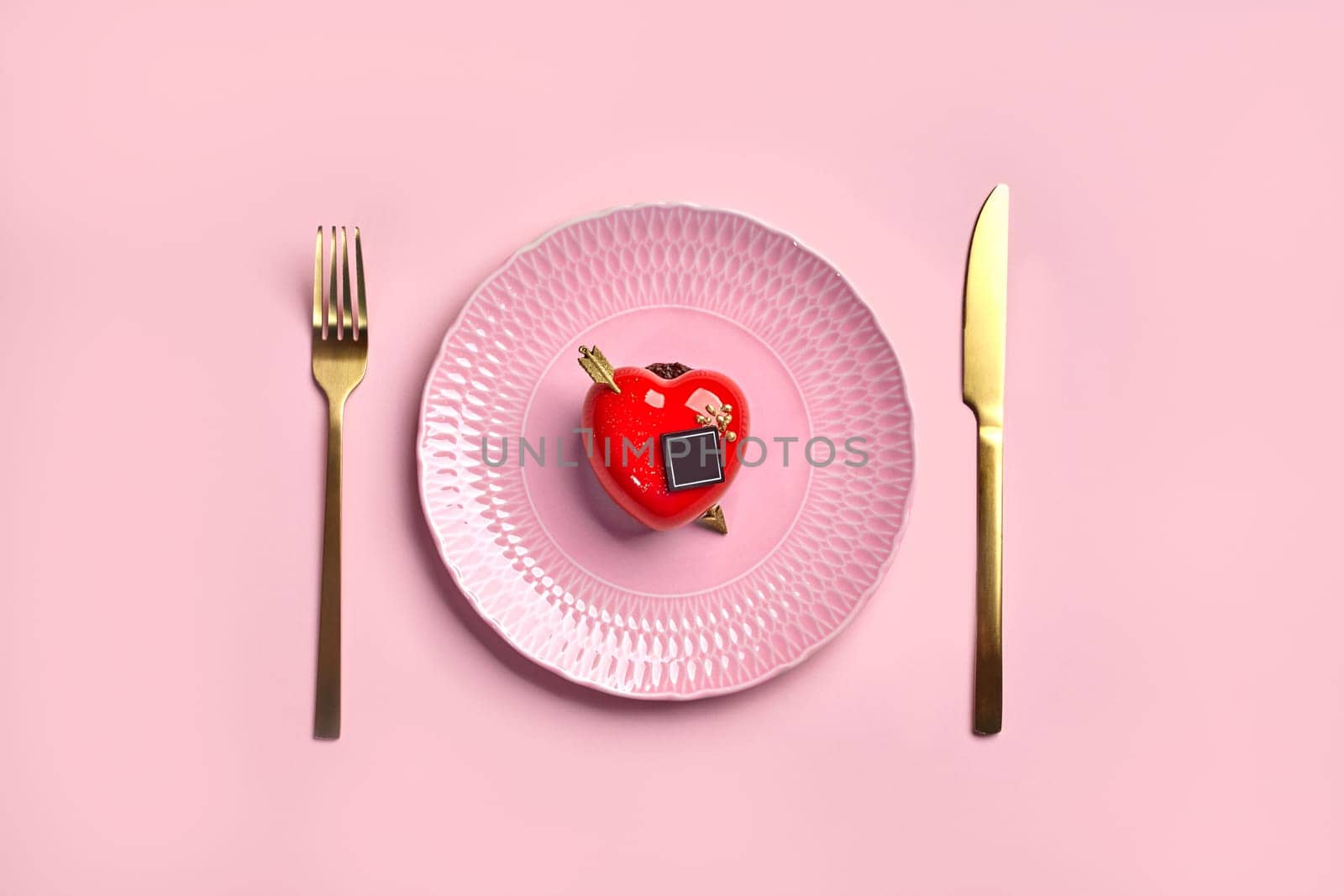 Red heart-shaped strawberry mousse dessert with glossy icing, chocolate and golden arrow elegantly placed on textured porcelain plate with gold cutlery on pink background. Romantic declaration of love