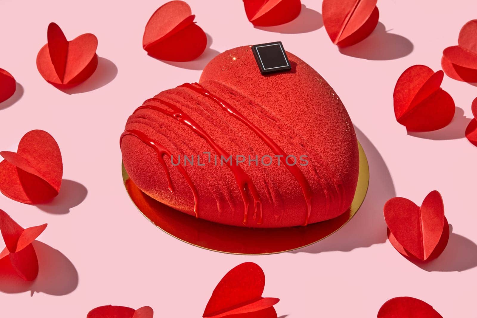 Bold red velvet heart-shaped mousse cake, presented on golden platform, amidst scattered paper hearts on delicate pink backdrop. Perfect for romantic celebrations and love-filled occasions