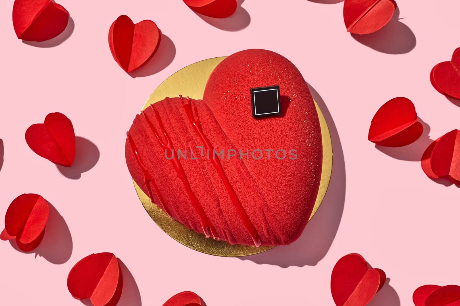 Vibrant heart-shaped red velvet cake on golden cardboard, surrounded by paper hearts on pastel pink background. Romantic dessert for declaration of love
