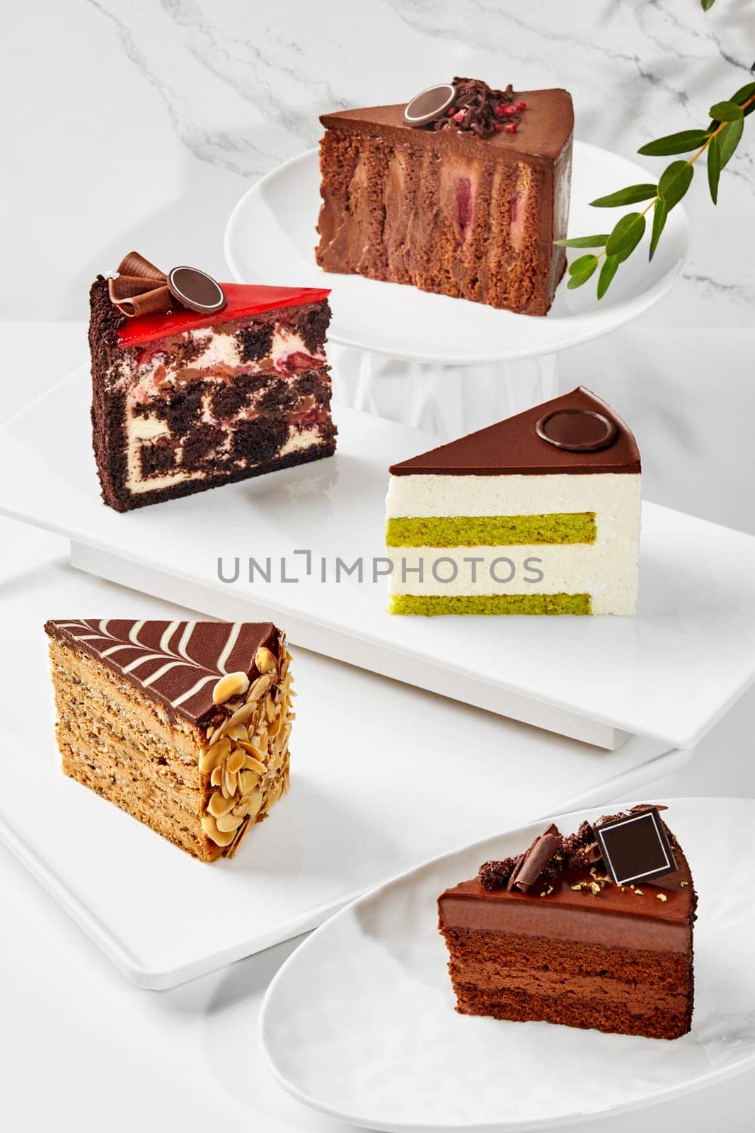 Tempting slices of various cakes with velvety chocolate layers, airy pistachio sponge, creamy mousse, nuts and berries arranged on white plates on marble surface. Patisserie masterpieces