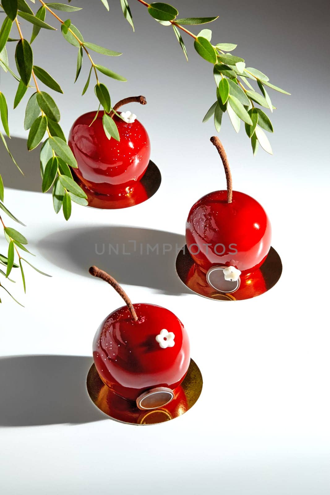 Evocative of cherry orchard, artisanal pastries with red glossy glaze and chocolate stems displayed on reflective gold cardboards under green tree branches. Artisan desserts concept