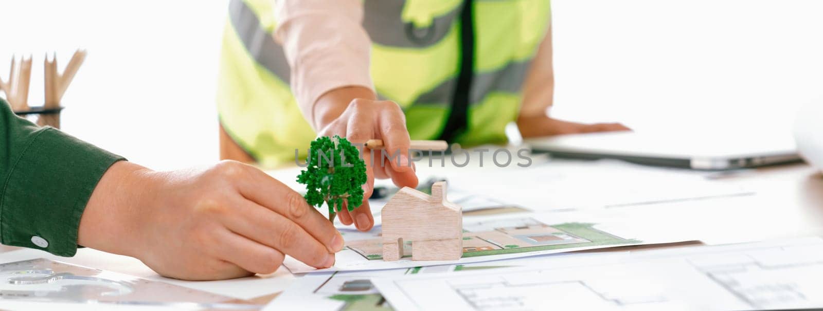 Professional architect and engineer collaborate to design eco-friendly house on meeting table with blueprint, wooden house block and tree model scatter around. Closeup. Delineation.