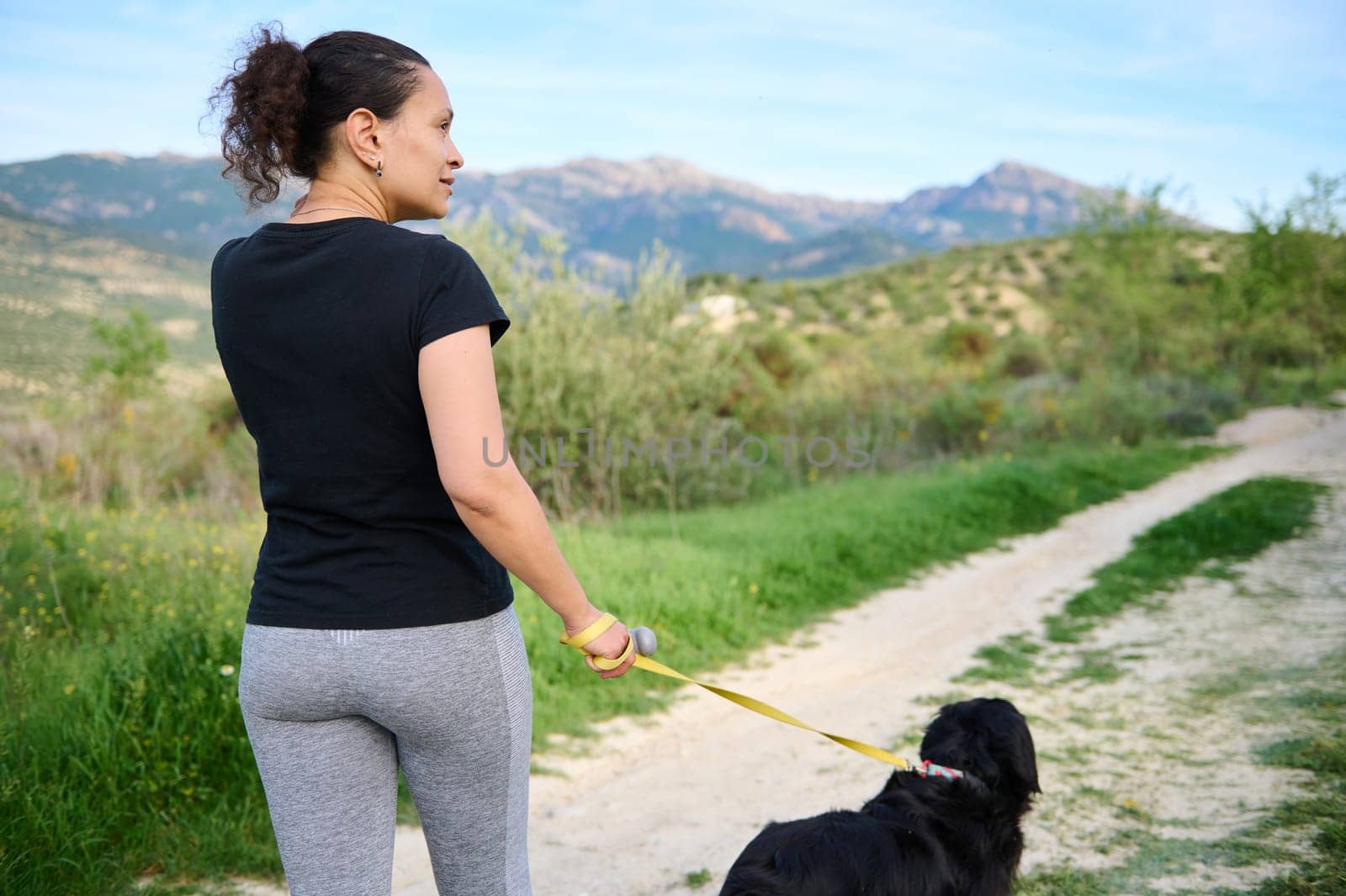 View from the back of a young sporty woman in gray leggings and black t-shirt, taking her dog for a walk while jogging outdoors in mountains nature