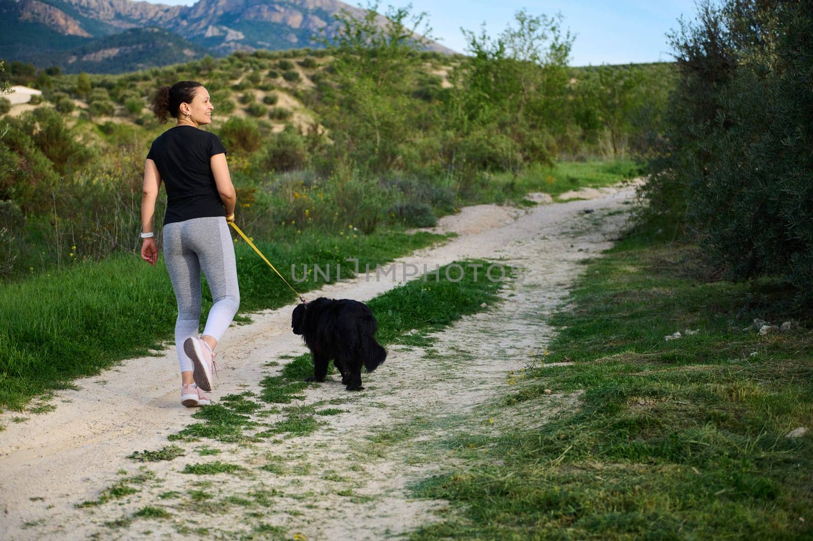 Full size shot of a happy woman smiling looking away, walking her dog in mountains outdoors. People, nature and playing pets concept. Rear view