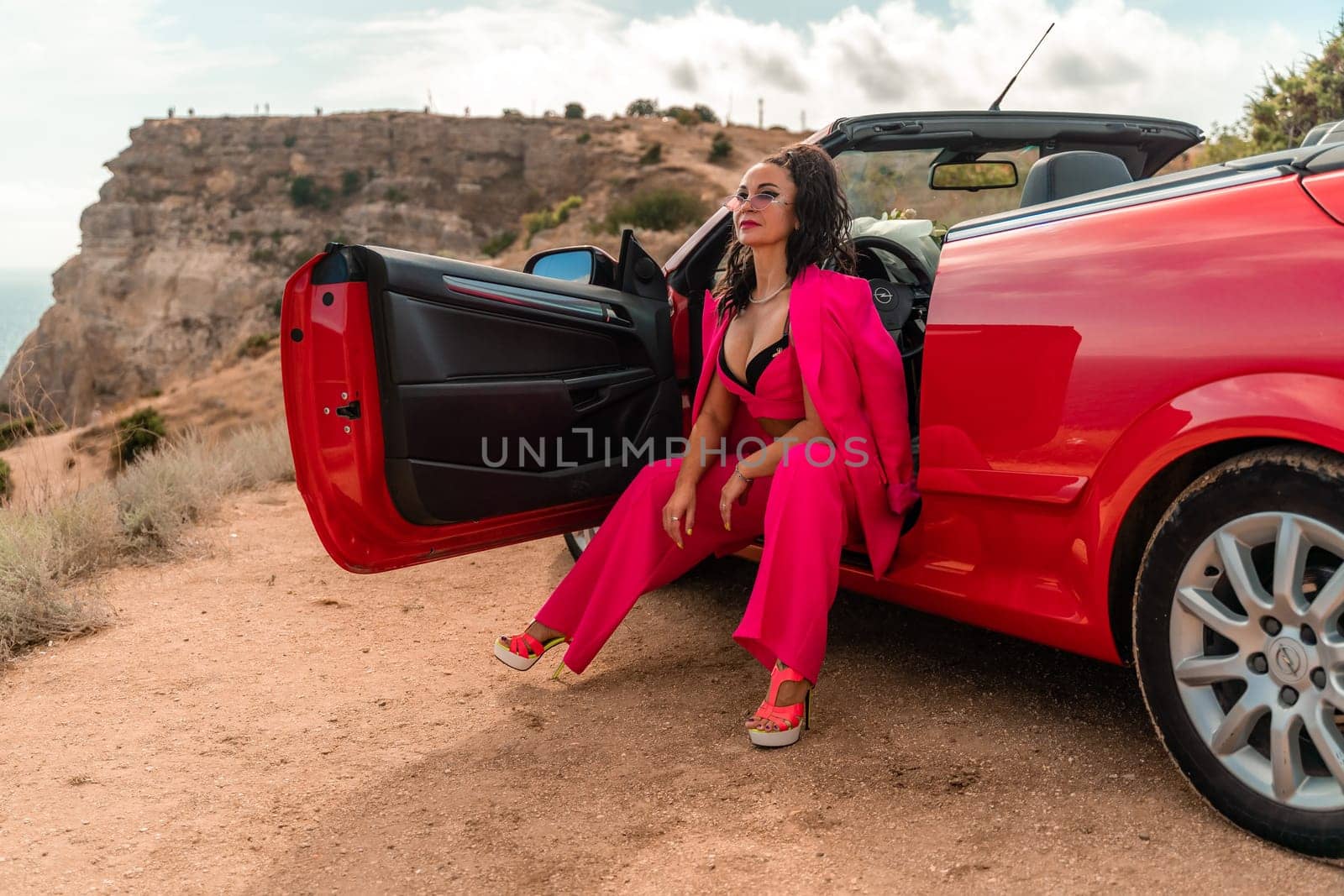 A woman in a pink suit sits in the open door of a red convertible. The scene is set on a rocky hillside overlooking the ocean. The woman is posing for a photo