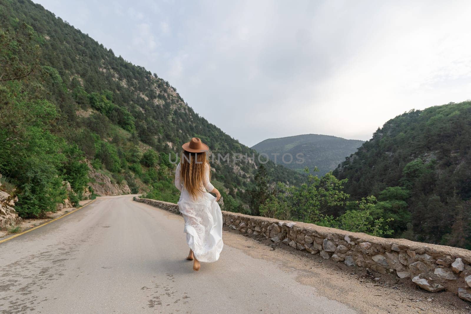 A woman in a white dress is walking down a road with a hat on. The road is surrounded by mountains and the sky is cloudy. The scene has a peaceful and serene mood