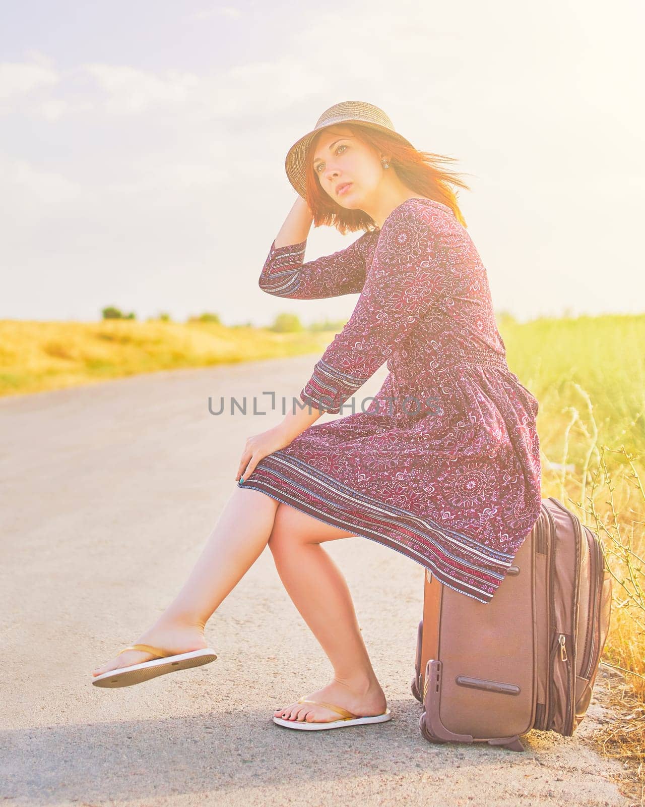 Amazing woman in summer dress and hat hitchhiking with suitcase on the road. Cute female sitting on Luggage waiting for car. Trip concept