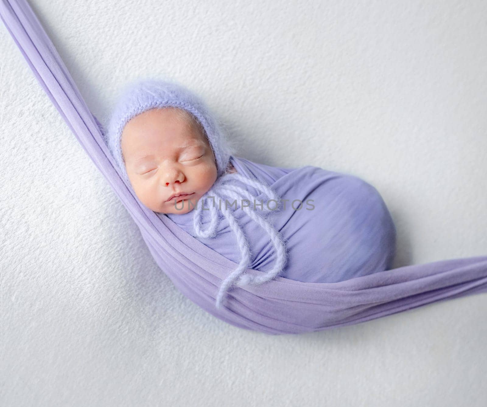 Newborn Baby In Lilac Wrap Sleeps In Hammock During Studio Photoshoot